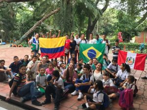 Intercambistas da AIESEC reunidos com várias crianças, sentados no chão e segurando bandeiras do Brasil, Peru e Chile, na Colômbia